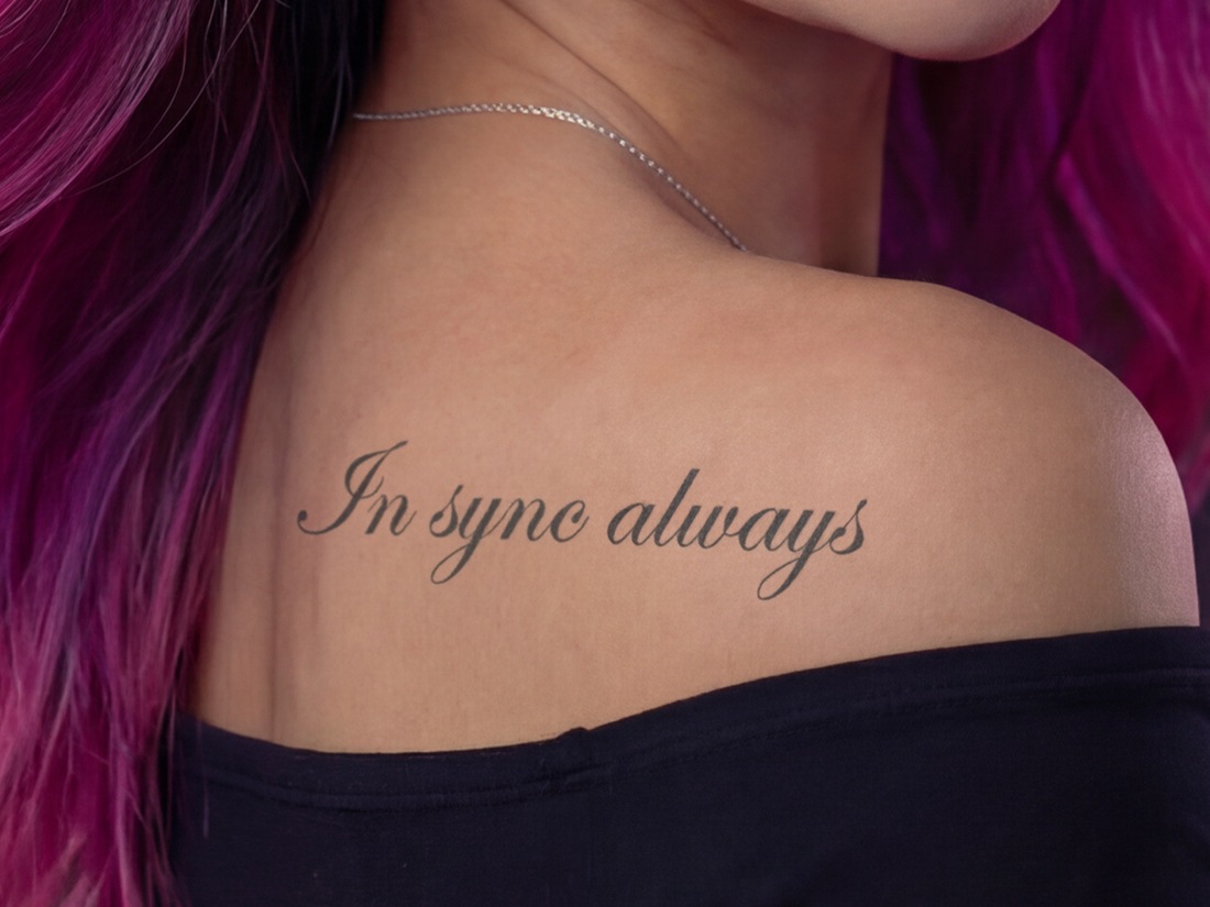 Vela Raye's right shoulder blade tattoo reading In sync always in Edwardian Script